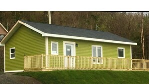 A single-story house with a green exterior, a dark roof, and a wooden deck. - Single Bedroom Green Bungalow (Rocky Harbour)