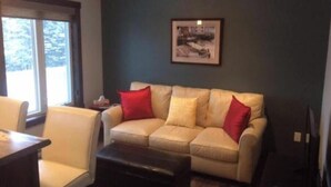 A living room with a beige sofa, red cushions, a dark coffee table, and a framed picture on the wall. - Single Bedroom Green Bungalow (Rocky Harbour)