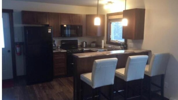 A modern kitchen with wooden cabinets, a black refrigerator, a microwave, and a stove. There are three bar stools with white covers. - Single Bedroom Green Bungalow (Rocky Harbour)