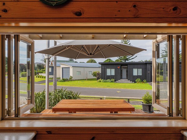 Vista desde una ventana a un deck de madera con una mesa y una sombrilla, con vista a un edificio moderno que tiene un garaje y un césped bien cuidado.