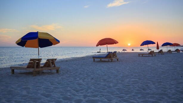 A beach with wooden lounge chairs and umbrellas, a sunset in the background, and calm sea.