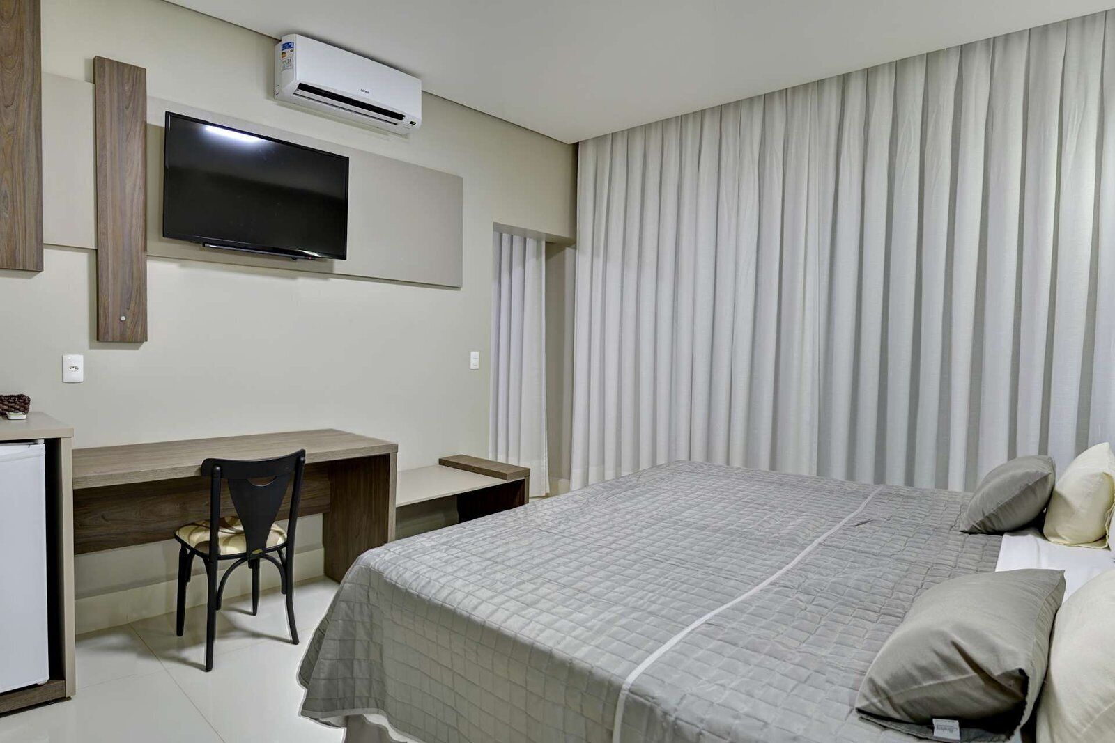 A hotel room with a bed, a desk, a chair, a television, and an air conditioning unit.