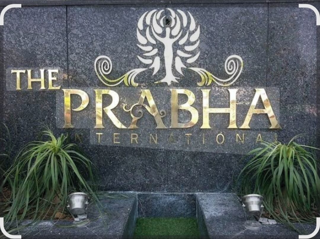 A stone wall with the text "THE PRABHA INTERNATIONAL" and a tree logo. There are two potted plants on either side of the wall.