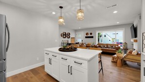 Una cocina amplia con armarios blancos y una isla central, un área de estar con sofá y mesa de centro, y un comedor con una mesa de madera y sillas.