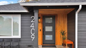 Una casa moderna con fachada gris oscuro, una puerta principal negra con paneles de vidrio y el número 2409 en la pared. Hay dos bancos de madera afuera y una pequeña planta en maceta cerca de la puerta.