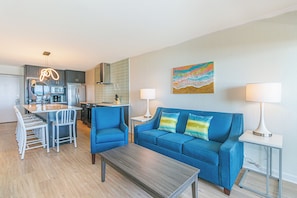 A modern living room with a blue sofa, a wooden coffee table, and a kitchen area with a refrigerator, microwave, and dining set.