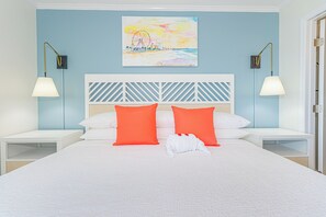 Une chambre d’hôtel avec un grand lit, deux tables de chevet, une tête de lit blanche et un tableau représentant une scène de plage.