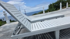 Ein weißer Lounge-Stuhl für den Außenbereich mit Metallgestell, auf einer Betonfläche platziert, mit Blick auf das Meer und viel Grün im Hintergrund.