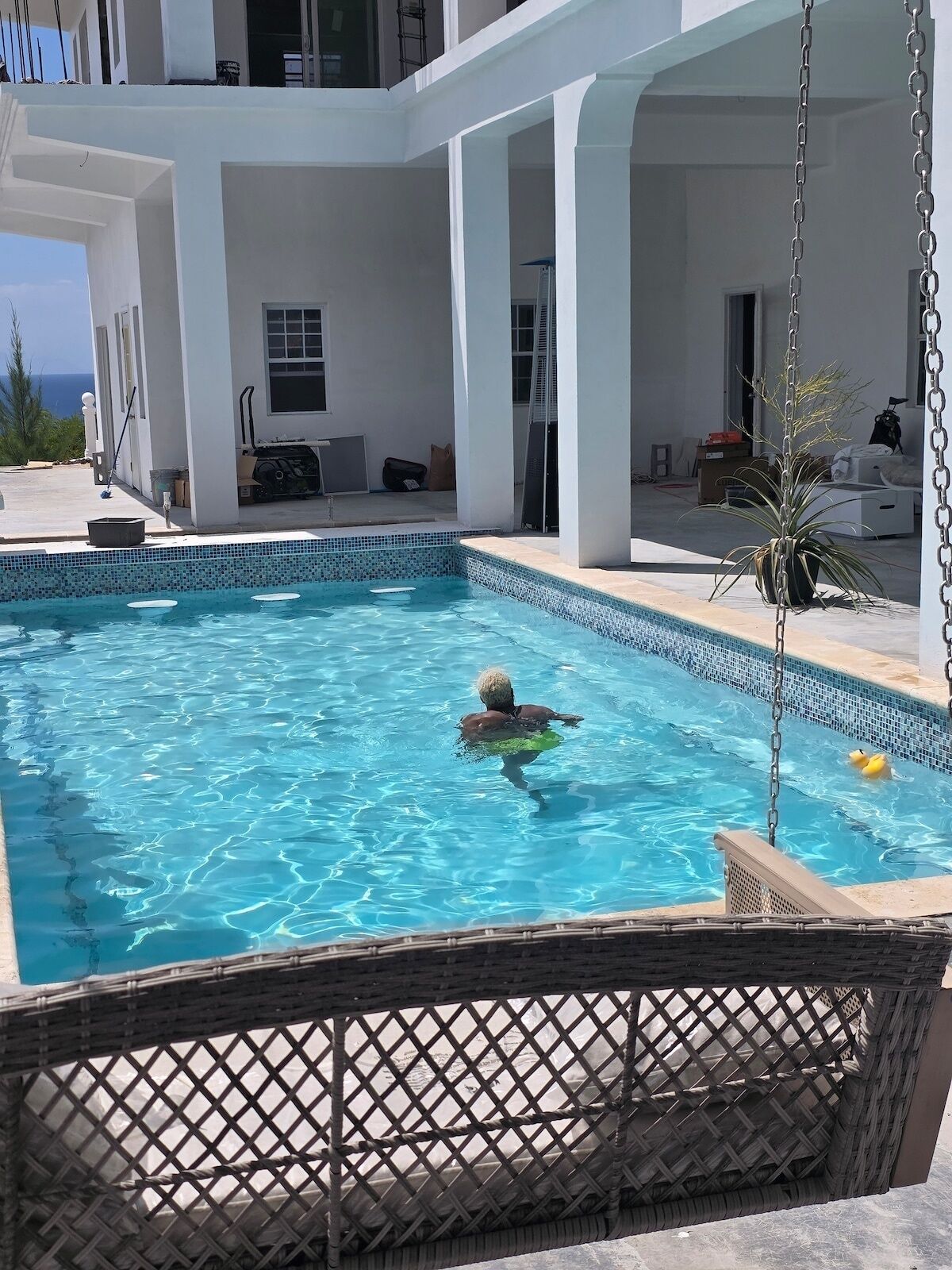 En swimmingpool med en person, omgivet af en hvid bygning og en overdækket patio.