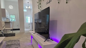 Ein modernes Wohnzimmer mit einem Flachbildfernseher auf einer weißen Konsole, einem grauen Shaggy-Teppich und einer Pflanze im Vordergrund.
