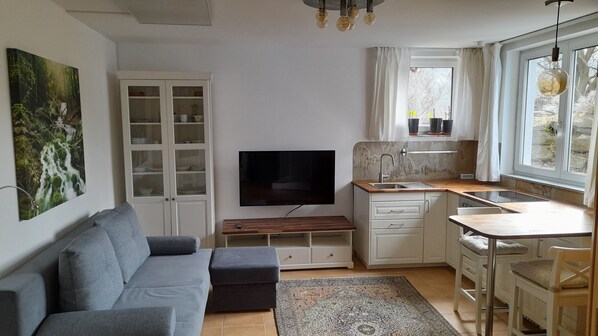 Een moderne woonkamer met een grijze bank, een flatscreen televisie en een eethoek met een kleine tafel en stoelen.