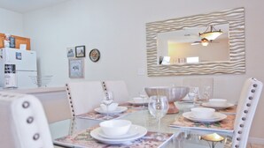 Un coin repas avec une table en verre dressée pour quatre personnes, entourée de chaises blanches à revêtement capitonné et agrémentées de clous en métal. Un miroir décoratif à cadre ondulé est accroché au-dessus de la table.