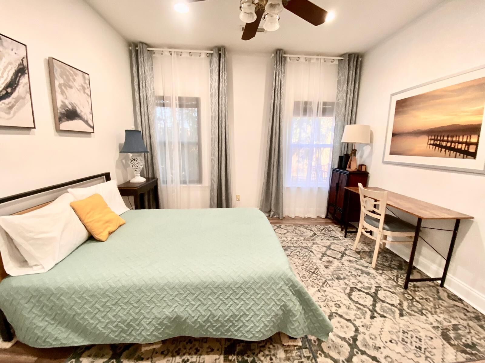 A bedroom with a bed, a desk, and a chair. There are two windows with curtains, a ceiling fan, and a painting on the wall.