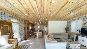 Ein großzügiger Wohnbereich mit einer großen Couch, einem Couchtisch und einem Fernseher. Es gibt eine Küchenzeile mit Holzschränken und einem Kühlschrank. Der Raum verfügt über eine Holzbalkendecke und Backsteinwände.