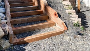 Eine Holztreppe führt zu einem Haus mit Stein Fundament, umgeben von einem Kiesweg und einem Holzzaun.