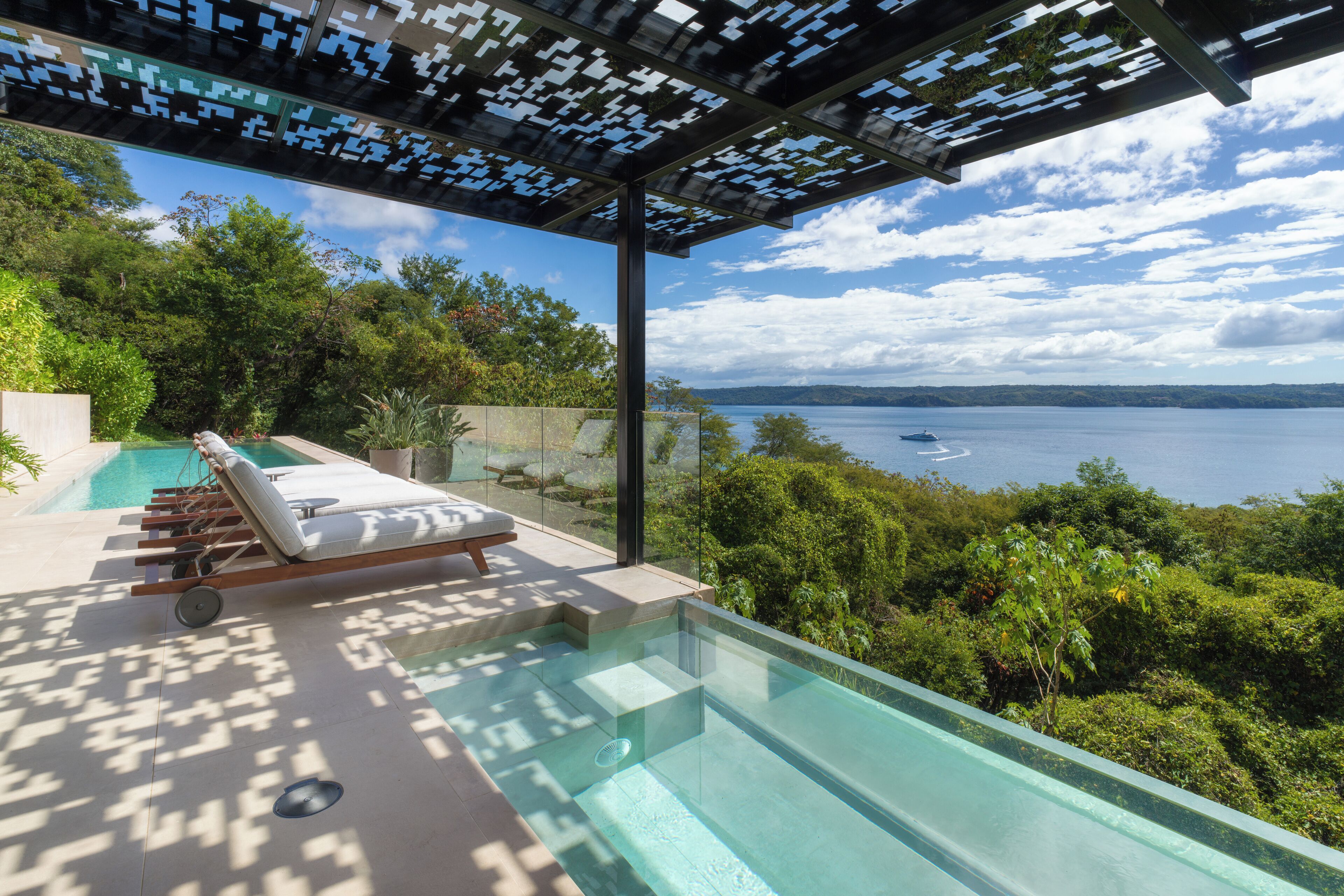 A glass-enclosed infinity pool with a lounge area, overlooking a body of water and lush greenery.