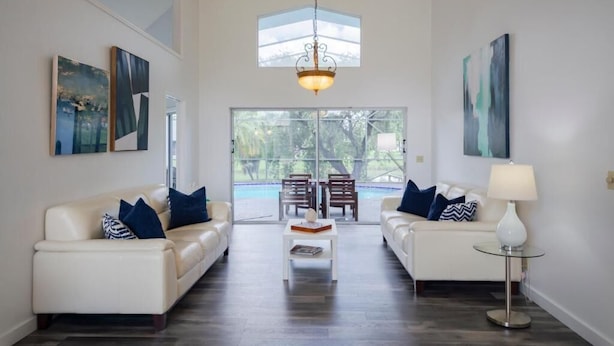 Un salon spacieux avec deux canapés blancs, une table basse et un lampadaire. Les murs sont ornés de tableaux abstraits et une grande fenêtre offre une vue sur les arbres.