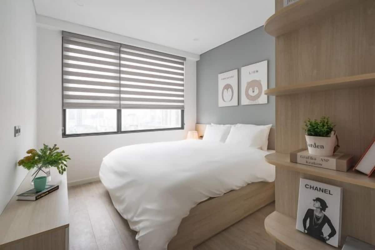 Un dormitorio moderno con una cama, un ventanal con persianas, una mesita de noche con una planta, y un estante con libros y una revista Chanel.