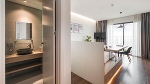 Ein modernes Hotelzimmer mit Holzboden, einer Essecke mit Tisch und Stühlen, einem großen Fenster mit Vorhängen und einem Badezimmer mit Waschbecken und Spiegel.