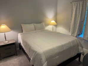 Una cama bien hecha con edredón blanco y almohadas, dos lámparas de noche, una mesita con cajón y una ventana con cortina.