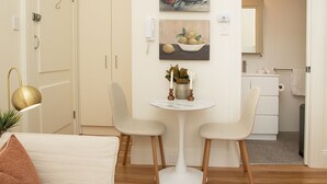 Quarto moderno e bem iluminado com área de jantar, destacando uma mesa redonda pequena, duas cadeiras e um vaso de planta. Há uma pintura na parede e um espelho sobre a pia.