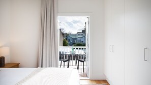 Ein Schlafzimmer mit einem Bett, einem Nachttisch, einer Lampe, einem Fenster mit Vorhängen und einem Balkon mit Stühlen und einem Tisch.