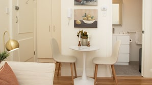 Quarto moderno e bem iluminado com área de jantar, destacando uma mesa redonda pequena, duas cadeiras e um vaso de planta. Há uma pintura na parede e um espelho sobre a pia.