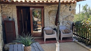 Un edificio de piedra con techo de tejas, una mesa de madera con una maceta tejida y dos sillas blancas bajo una pérgola.