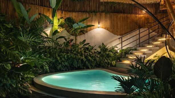 Une piscine entourée d’une végétation tropicale luxuriante, un escalier en bois et une structure surmontée d’un toit de chaume.