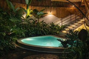 A pool surrounded by lush tropical plants, a wooden staircase, and a thatched roof structure.