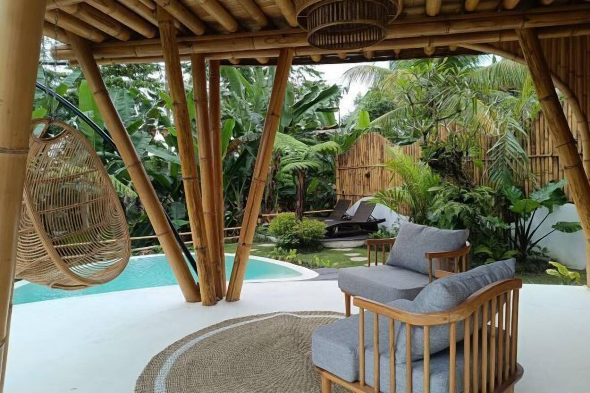Une pergola en bambou avec un fauteuil suspendu, une piscine et un espace détente avec des coussins.