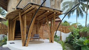 Une structure en bambou comprenant un espace salon et un fauteuil suspendu, le tout entouré d’une végétation luxuriante.