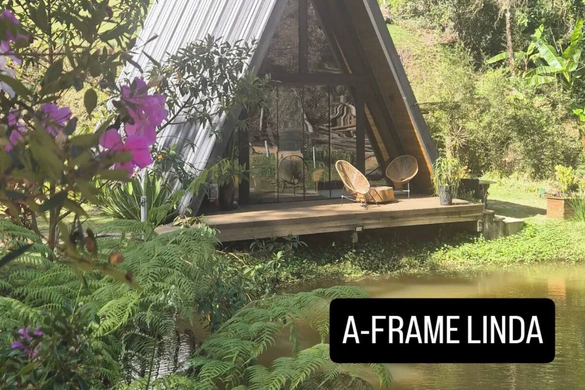 A small, triangular wooden structure with a deck, surrounded by lush greenery and a pond.