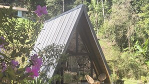 A small, triangular wooden structure with a glass enclosure, situated by a calm pond surrounded by lush greenery and flowering plants. - SEU PARAISO NA MONTANHAS (Marechal Floriano)