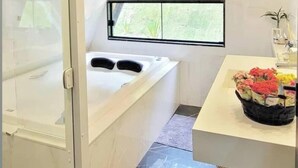 Un baño moderno con cabina de ducha de vidrio, una bañera independiente, un ventanal con vistas a zonas verdes y una pequeña mesa con un arreglo floral.