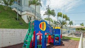 Un parque infantil con estructuras de juego coloridas, que incluyen un tobogán y elementos para escalar, junto a un edificio con una escalera y una colina cubierta de césped.