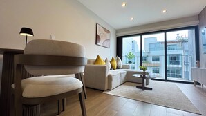 A modern living room with a sofa, a chair, a small table, and a floor lamp. There is a large window with a view of buildings outside. - Luxury Stay Miraflores | Entre Marriott & Pullman (Miraflores)