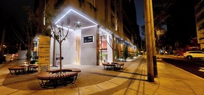 A hotel entrance with a modern design, featuring a reception desk, a signboard, and a seating area with tables and chairs.