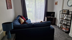 A living room with a dark blue sofa, a flat-screen TV, a bookshelf, and a window with curtains. - Spacious 4BR Home w/ Parking – Family-Friendly (Birstall)