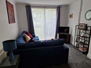 Een woonkamer met een donkerblauwe bank, een flatscreen televisie, een boekenkast en een raam met gordijnen.