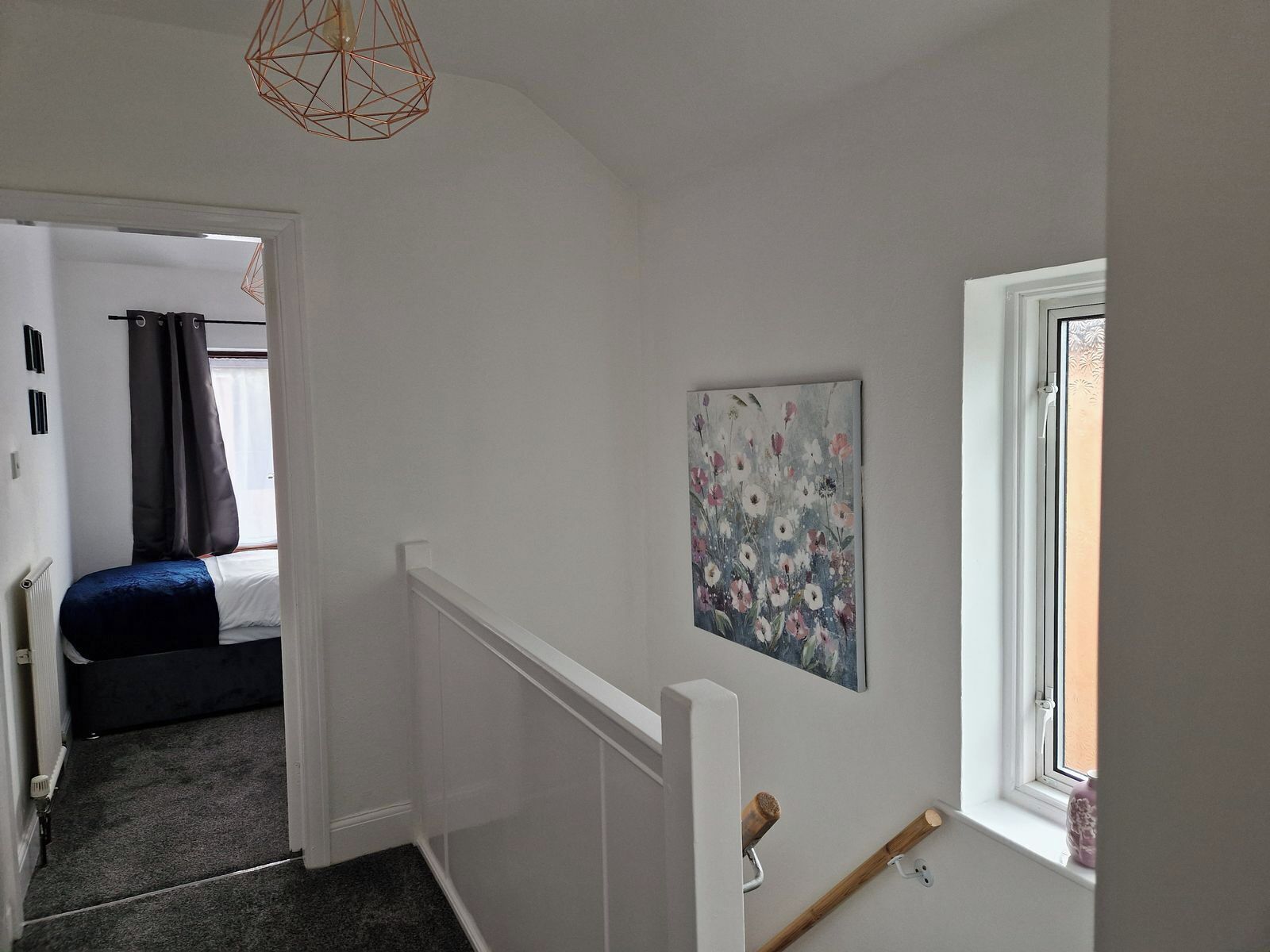 A narrow hallway with a bed in the background, a painting on the wall, and a window with a view.