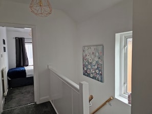 Een smalle gang met een bed op de achtergrond, een schilderij aan de muur en een raam met uitzicht.