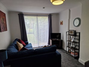 A living room with a dark blue sofa, a television on a stand, a bookshelf, and a large window with curtains.