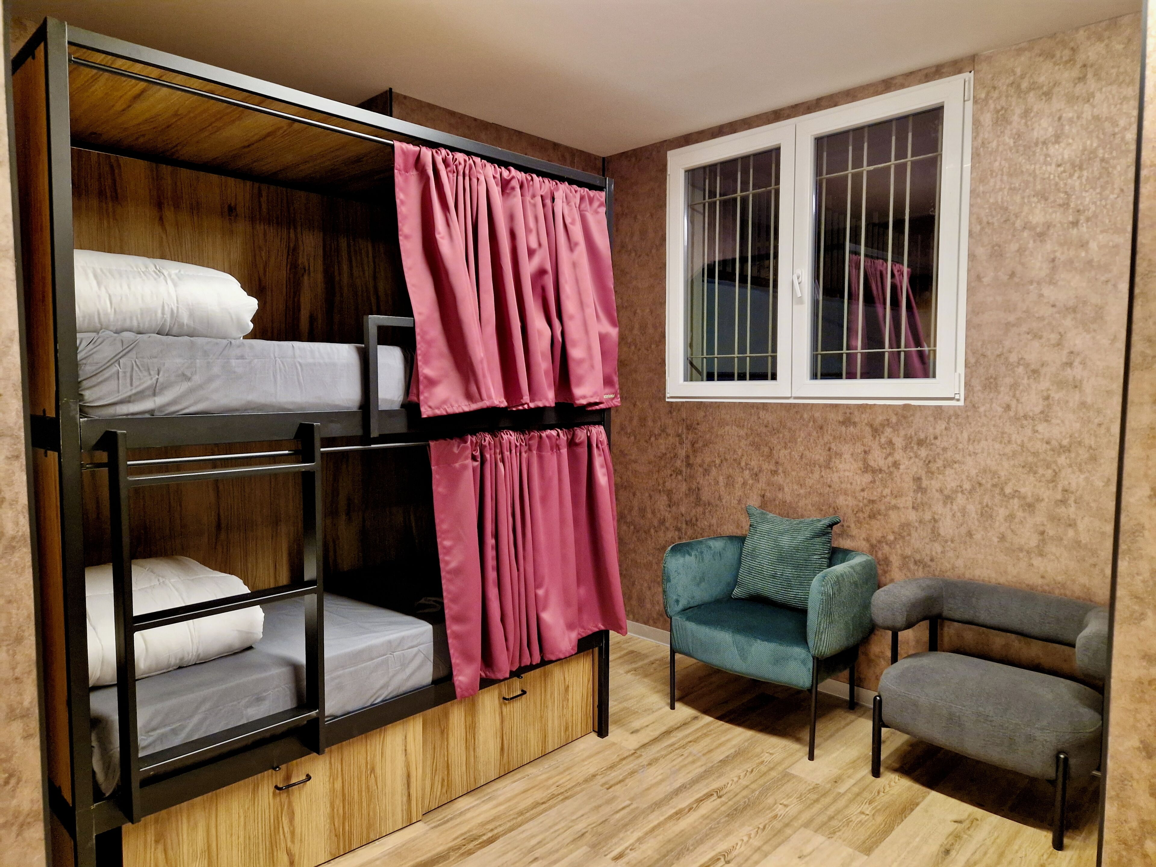 A dorm-style room with bunk beds, a window with bars, a chair, and a small table.