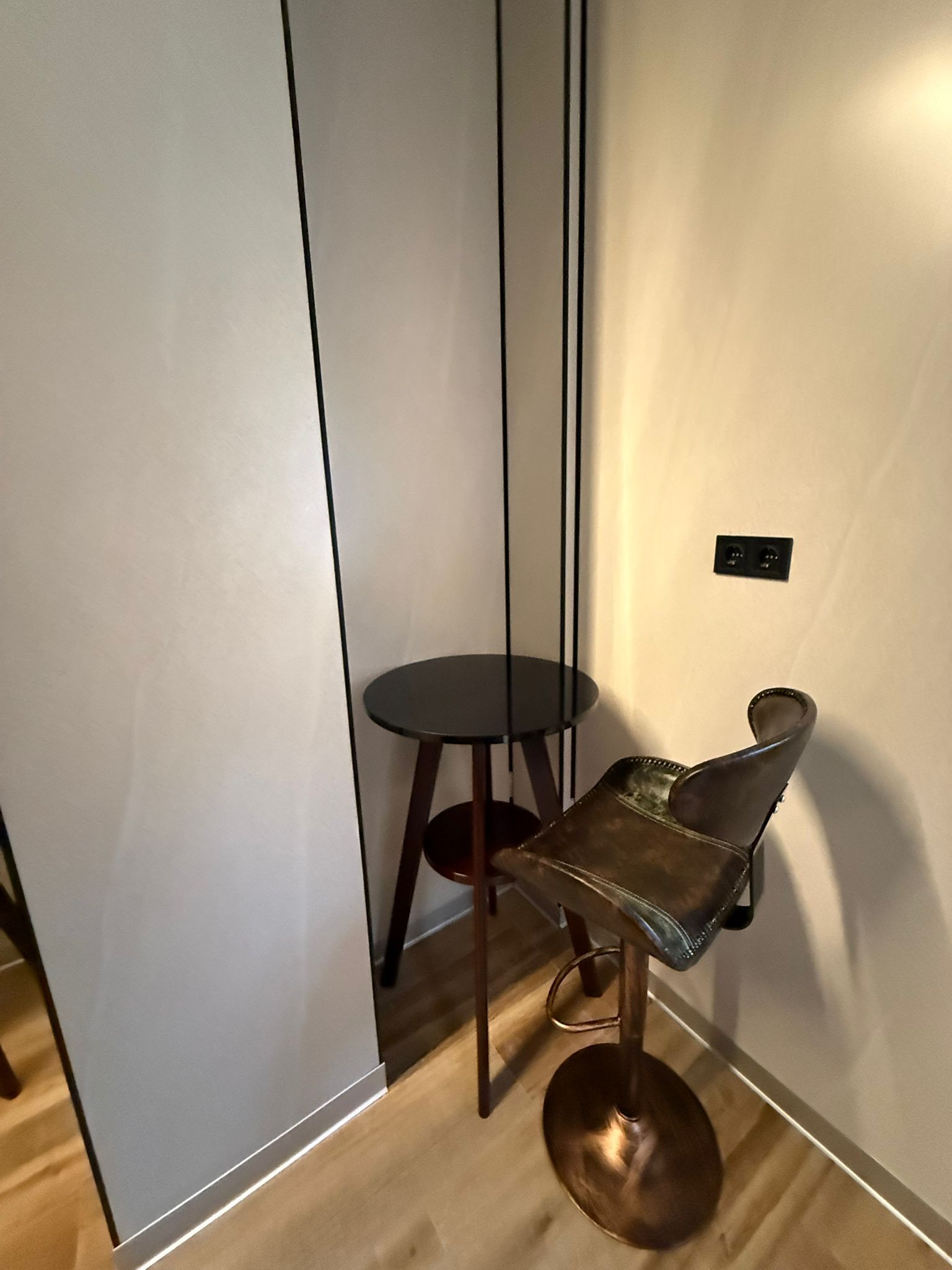 Un rincón de una habitación con piso de madera, una mesa redonda pequeña y una silla de base metálica.