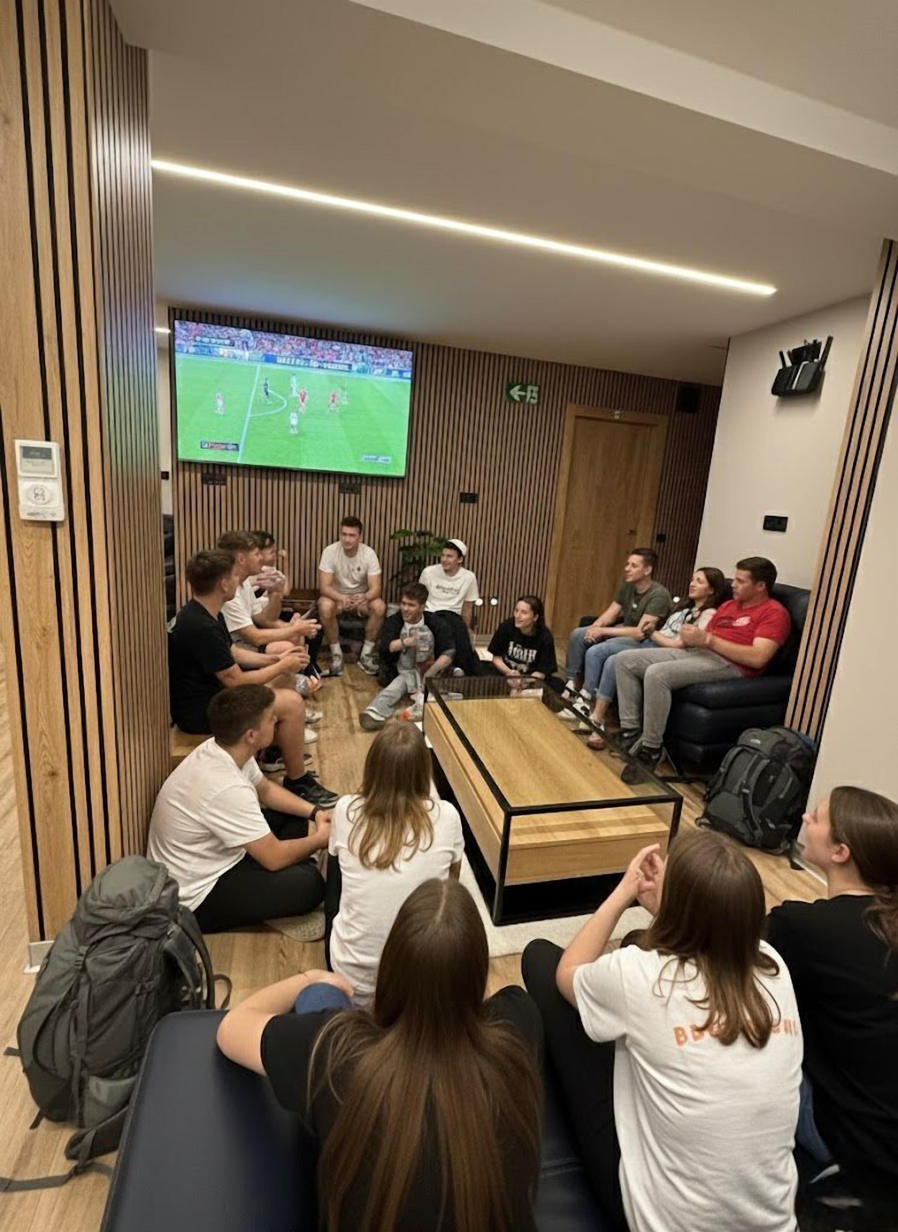 A group of people seated in a modern lounge area with a large TV screen displaying a sports event, wooden paneling on the walls, and a coffee table in the center.