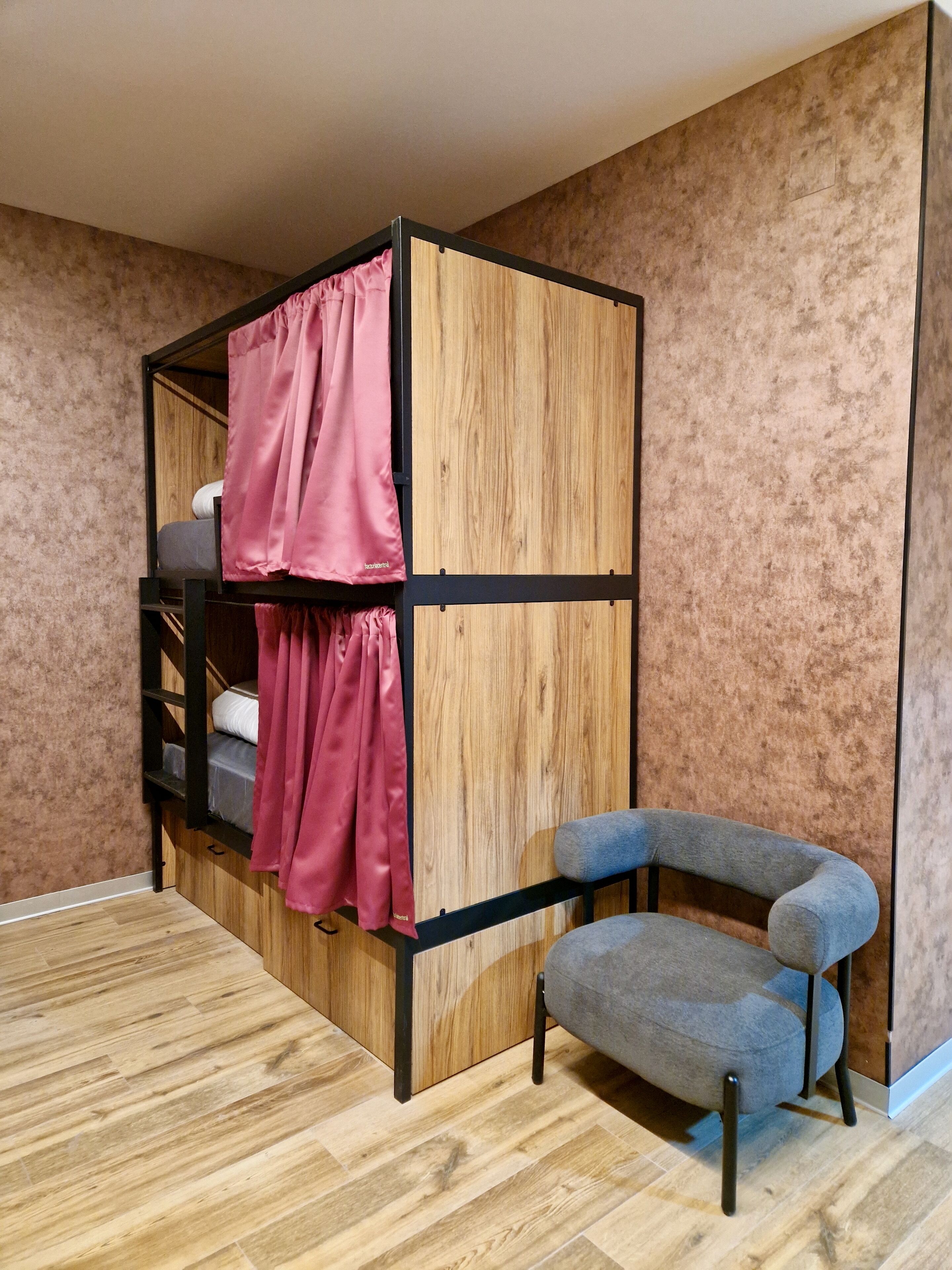 A bunk bed with pink curtains, a gray chair, and wooden flooring.