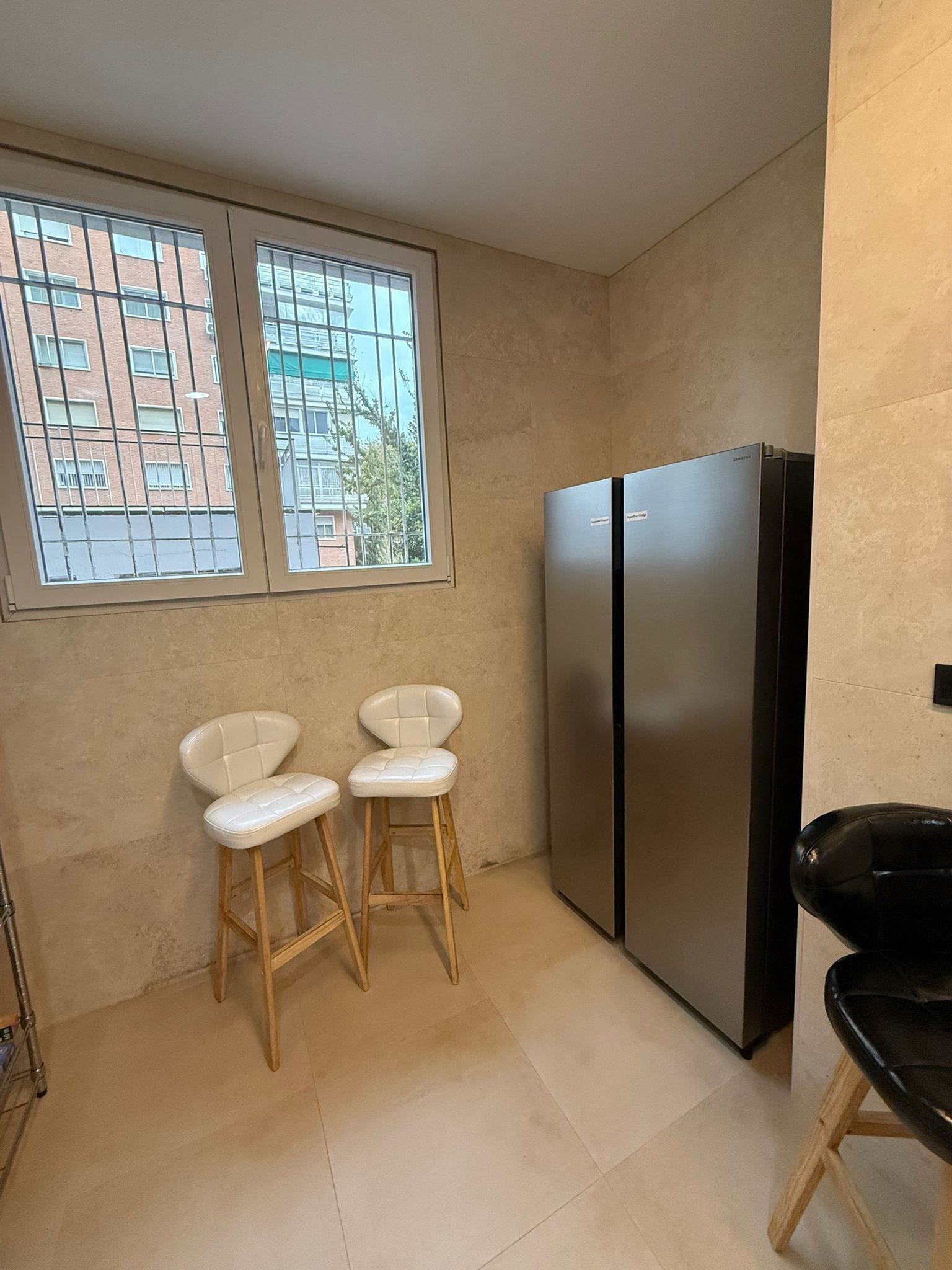 A small kitchen with two white bar stools, a black refrigerator, and a window with a view of buildings.