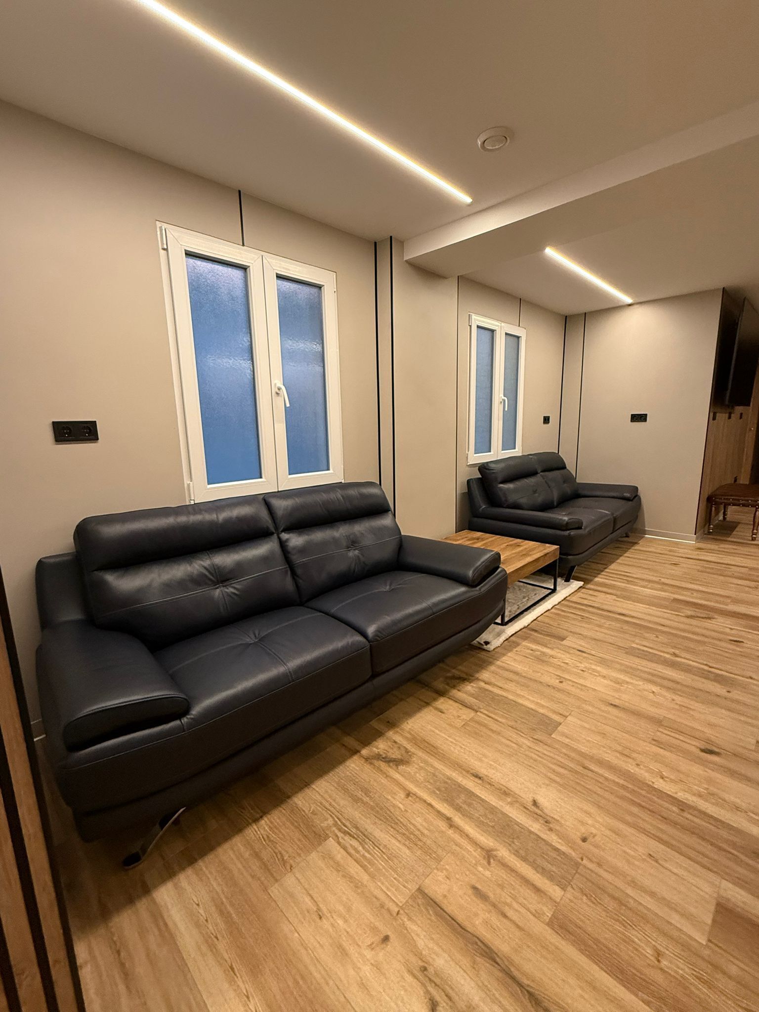A modern living room with a black leather sofa, wooden flooring, and built-in windows with frosted glass.
