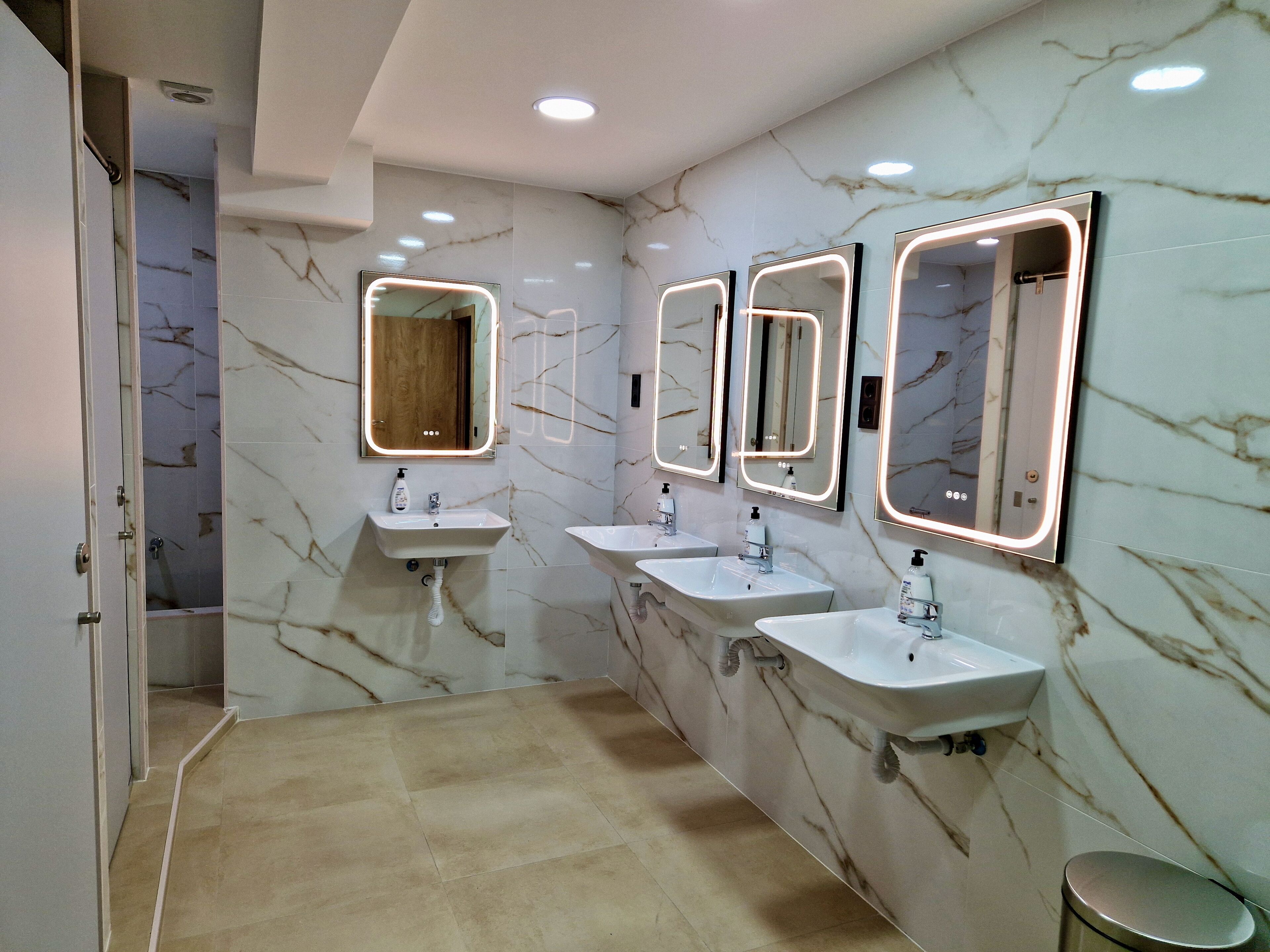 A modern bathroom with three sinks, marble walls, and illuminated mirrors.